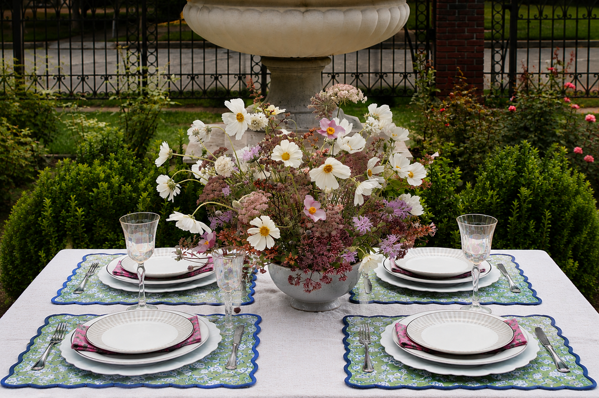 An outdoor dining table set with four white plates, clear wine glasses, and pink napkins on blue and green patterned placemats, centered with a large flower arrangement in a white pot. The background shows a garden with green hedges, a stone fountain, and a black metal fence.