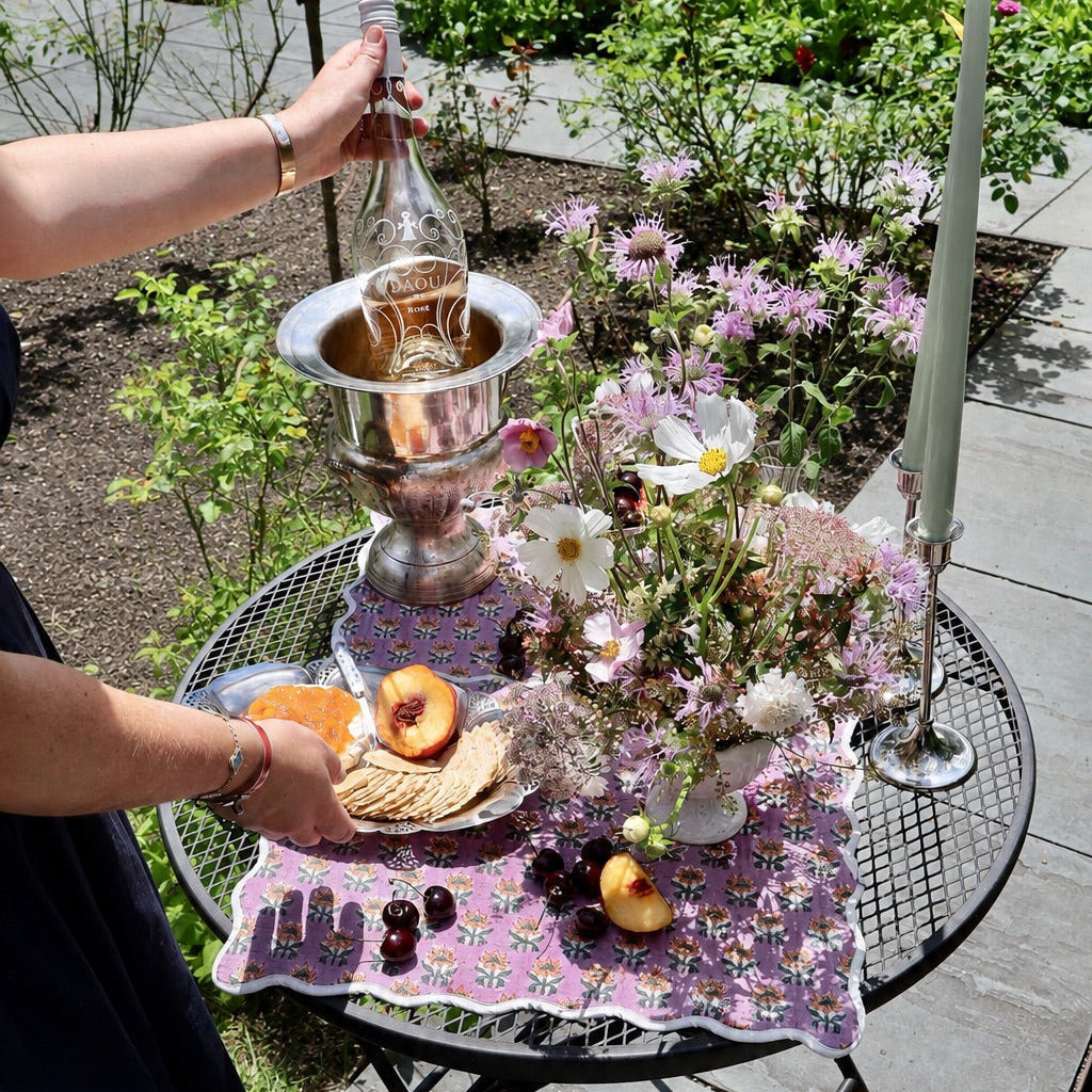 A lifestyle shot of a woman pouring champagne from a bottle into a silver ice bucket on a black metal mesh outdoor table. The table is decorated with a purple floral tablecloth, a flower arrangement, crackers, fruit, and candles, set in a garden patio environment.