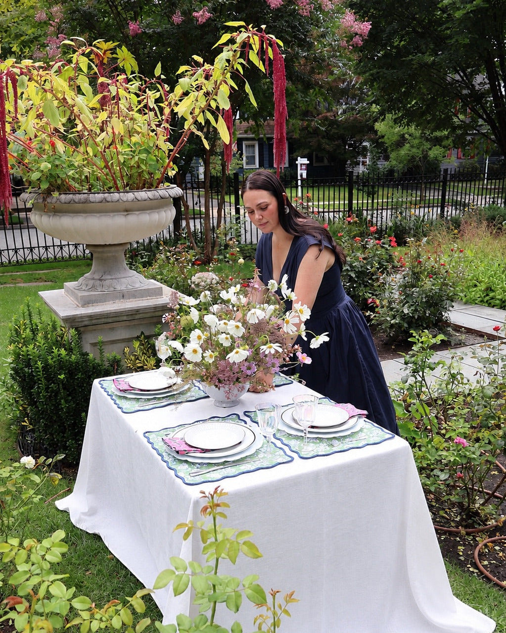 A woman in a navy blue dress stands beside an outdoor garden table draped with a white tablecloth, set with plates, pink napkins, and a floral centerpiece.