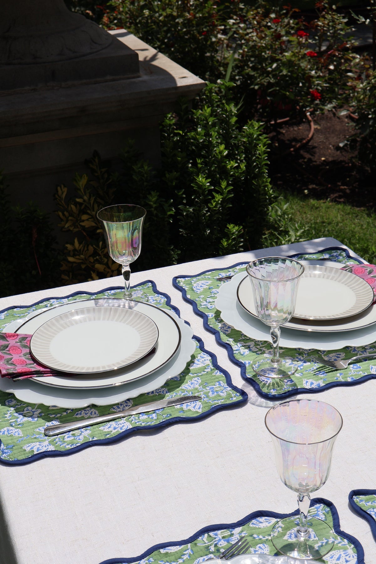 Outdoor table setting with patterned placemats, white plates, clear stemmed glasses, and a white tablecloth, photographed in a garden environment.