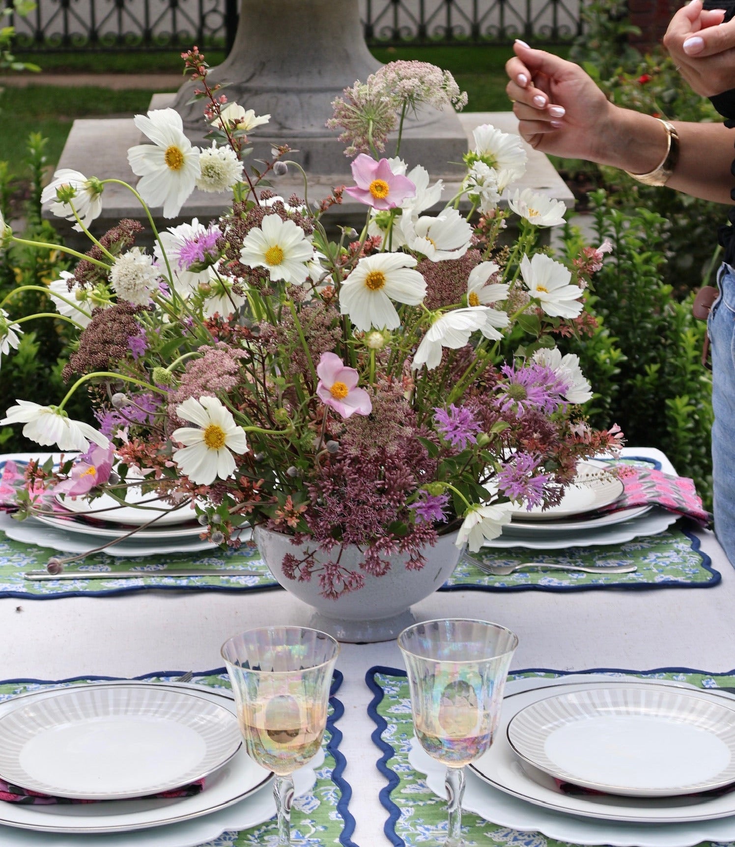 A styled outdoor dining table arrangement with a large floral centerpiece in a white bowl, white plates, patterned placemats, and two iridescent goblets. A person is partially visible on the right side in a garden setting with a stone fountain in the background.