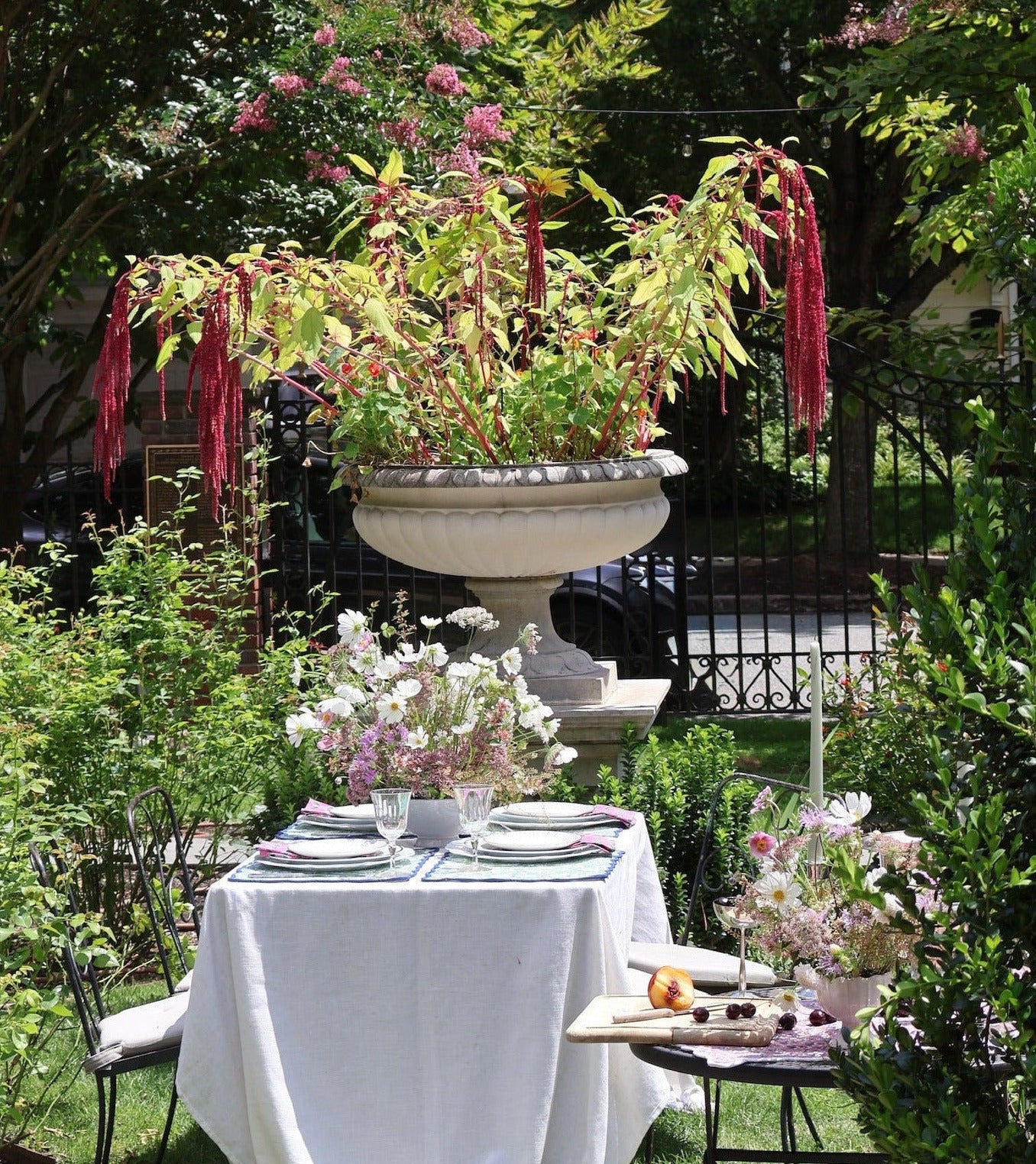 An outdoor garden table setup with a white draped tablecloth, floral centerpieces, plates, and chairs, surrounded by lush greenery, pink blossoms, and a large decorative urn planter.